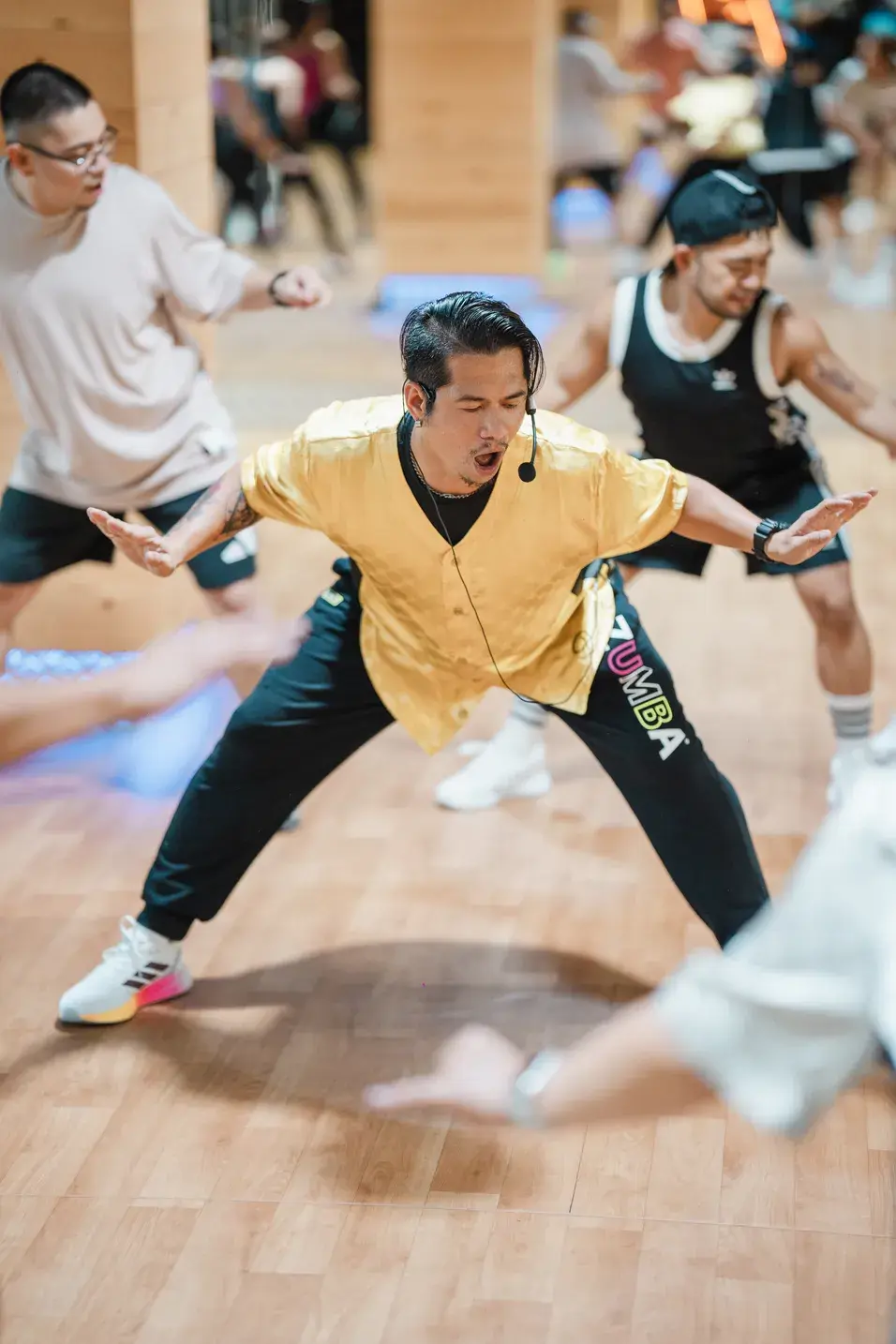 Energetic BODYJAM instructor in a yellow shirt leading a lively dance routine at GymNation, surrounded by motivated participants following his moves in a vibrant fitness studio.