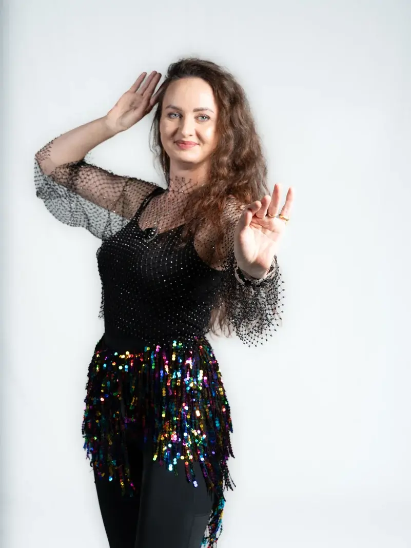 A professional belly dance instructor posing gracefully in a sparkling black outfit with sequined hip scarf, smiling confidently against a white background.